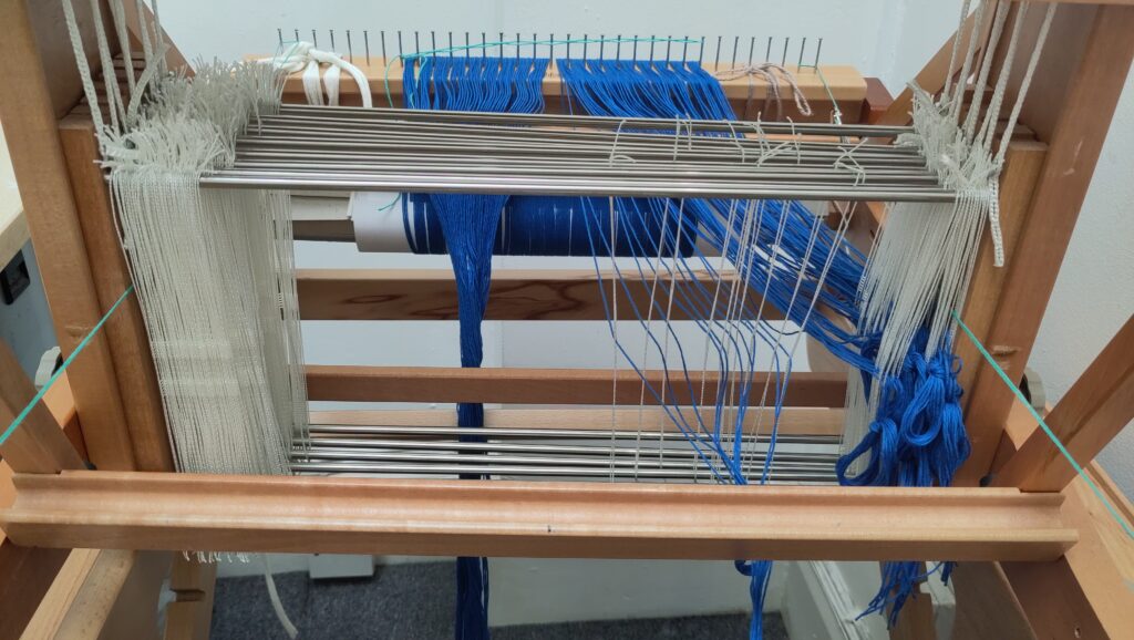 My loom in the process of being threaded. Ten string heddles have been selected in a pattern, with blue warp threads threaded through them.